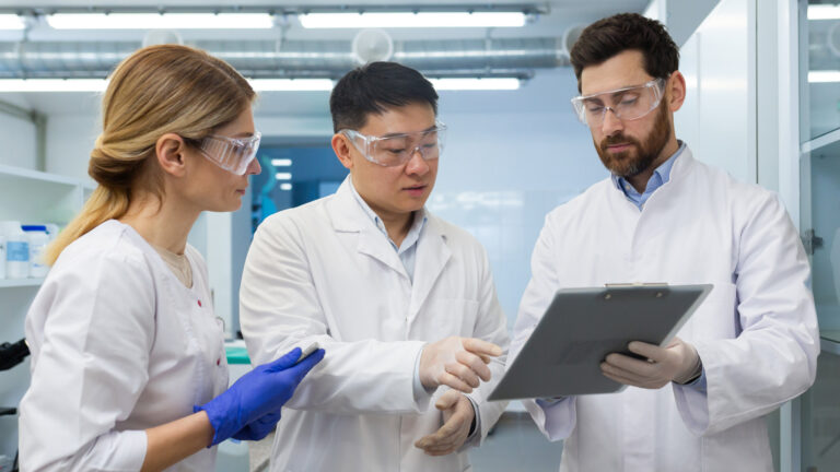 Group in labcoats and safety glasses pointing at clipboard