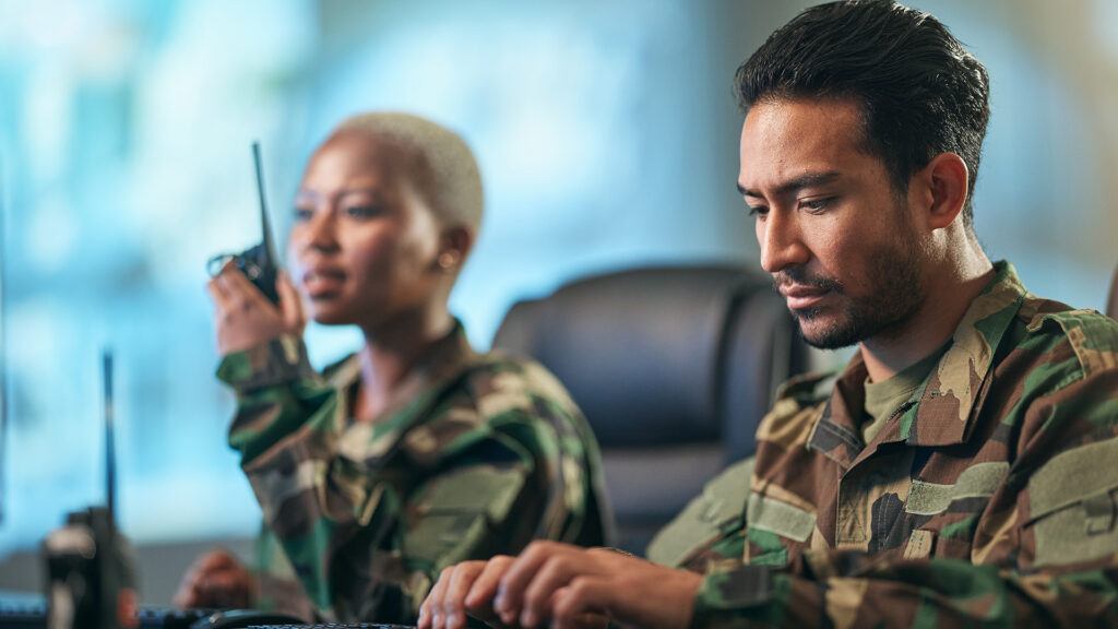 Man in military fatigues types on computer with woman fatigues on walkie talkie in background