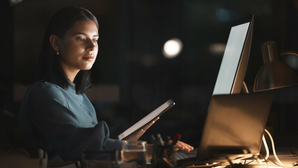 Young business woman in front of computer holding iPad at desk at night