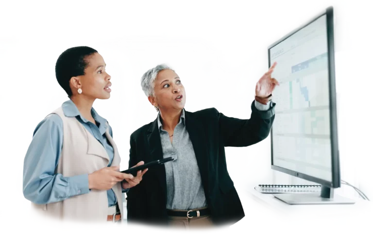 Older business woman pointing to large monitor for younger business woman holding iPad